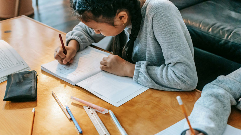 En flicka skriver i en anteckningsbok. På bordet ligger utspridda pennor.