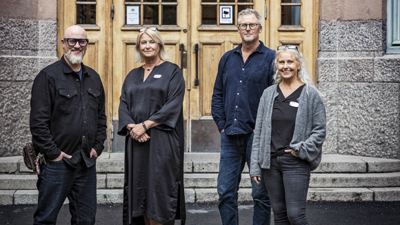Fyra personer står utanför en skolentré och tittar leende in i kameran.