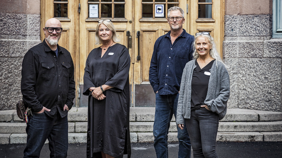 Fyra personer står utanför en skolentré och tittar leende in i kameran.
