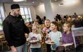 Kökschef tillsammans med glada skolbarn som hämtar mat i matsal.