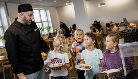 Kökschef tillsammans med glada skolbarn som hämtar mat i matsal.