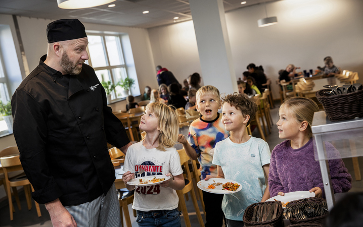 Kökschef tillsammans med glada skolbarn som hämtar mat i matsal.