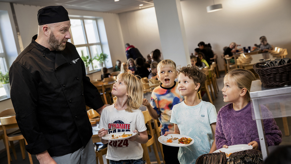 Kökschef tillsammans med glada skolbarn som hämtar mat i matsal.