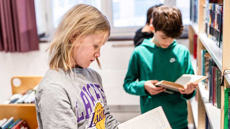 Två elever står framför en bokhylla i ett bibliotek och läser i varsin bok.