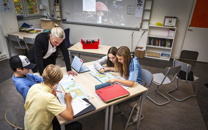 Klassrum med fyra ungdomar som arbetar med sina geografiböcker. Deras lärare ger stöd.