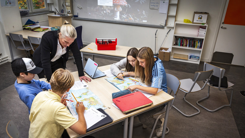 Klassrum med fyra ungdomar som arbetar med sina geografiböcker. Deras lärare ger stöd.