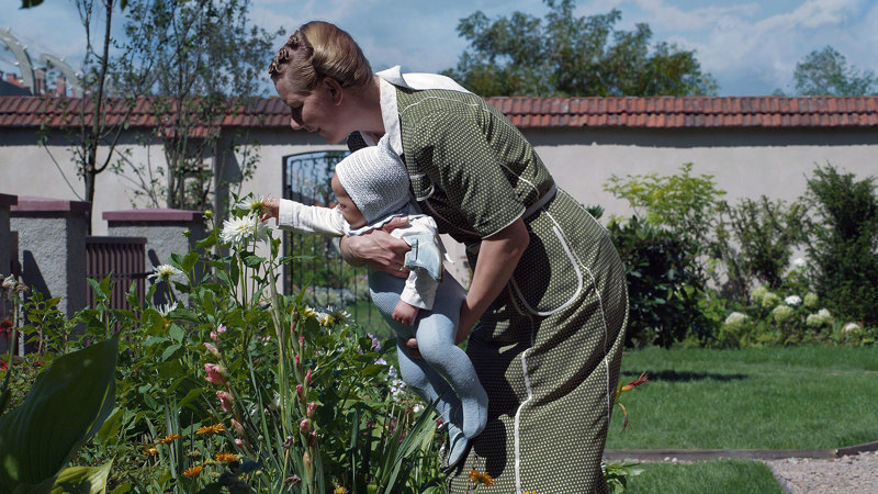 En kvinna står i en trädgård och håller en bebis i famnen som känner på en blomma.