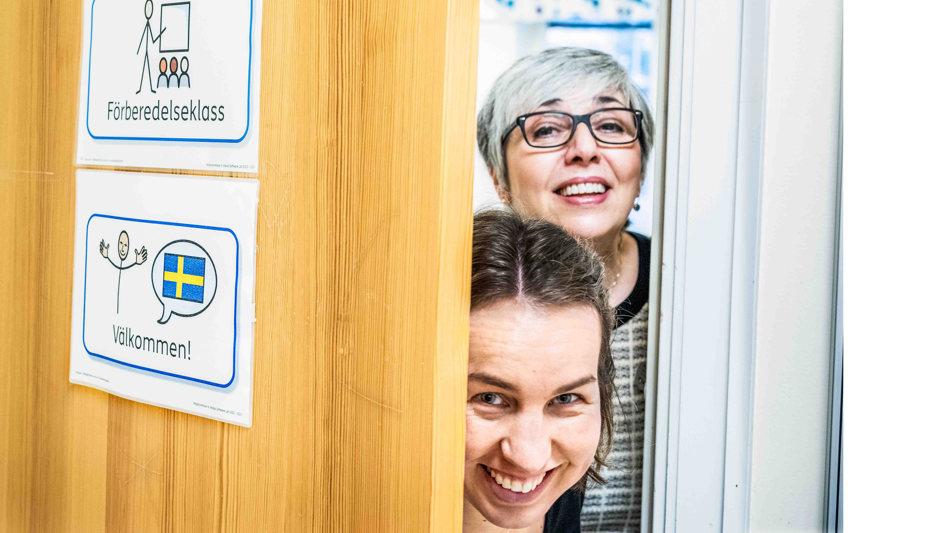 Två glada kvinnor tittar fram genom en dörröppning i ett klassrum. 