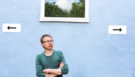En man står med armarna i kors framför en blå husvägg. 