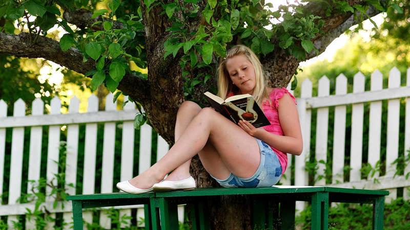 En flicka sitter på en bänk under ett träd och läser en bok.