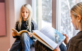 Två flickor sitter i ett fönster och läser.