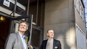 Två män står framför entrén till en byggnad. Över dem syns en skylt där det står 
