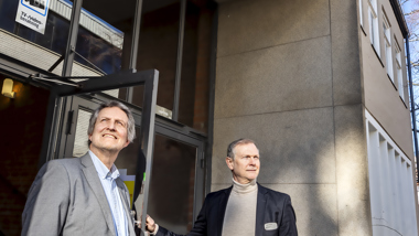 Två män står framför entrén till en byggnad. Över dem syns en skylt där det står 