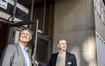 Två män står framför entrén till en byggnad. Över dem syns en skylt där det står 