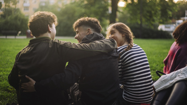 Tre unga personer sitter med ryggarna mot kameran på en gräsmatta, två håller armarna om varandra.