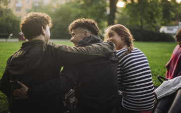 Tre unga personer sitter med ryggarna mot kameran på en gräsmatta, två håller armarna om varandra.