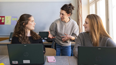 En lärare står och pratar med två elever i ett klassrum.