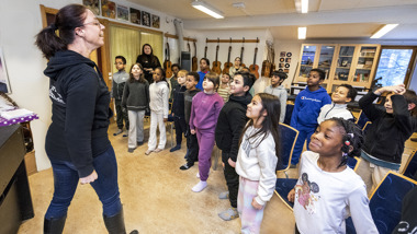 En klass med elever och en lärare står upp i ett klassrum.