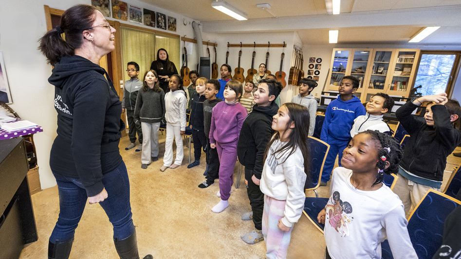 En klass med elever och en lärare står upp i ett klassrum.