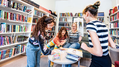 En lärare och tre elever i ett skolbibliotek. På ett bord ligger böcker och lappar med ord.