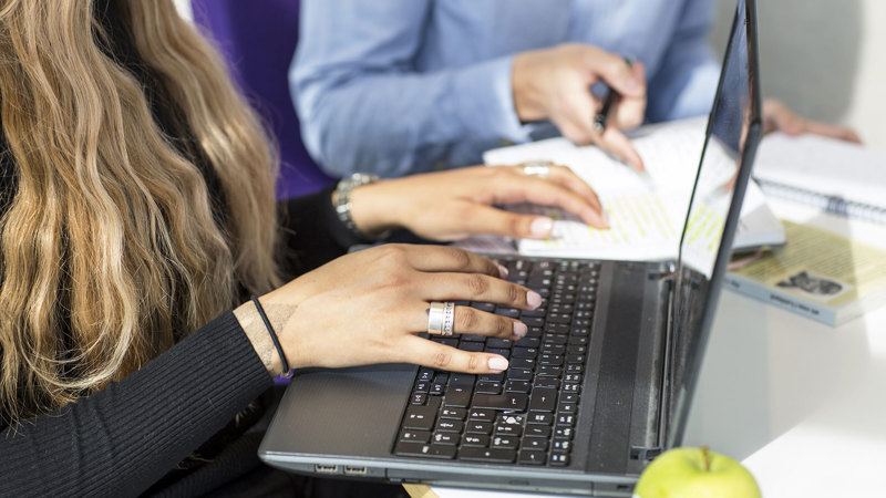 Två personer arbetar vid en bärbar dator  Den ena skriver på tangentbordet, den andra antecknar.