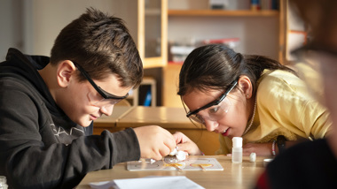 Två elever med skyddsglasögon tittar nära på ett experiment på bordet.
