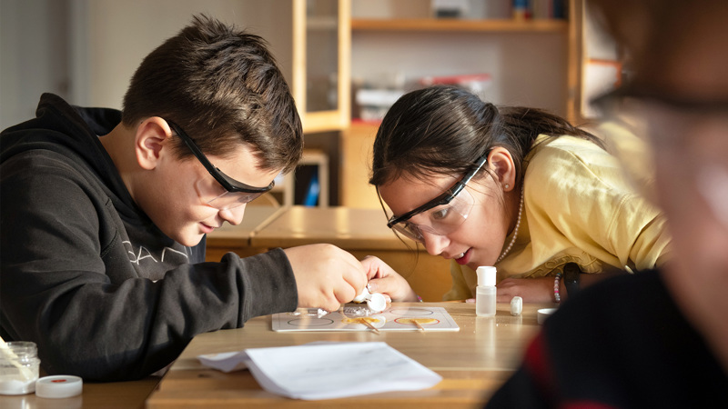 Två elever med skyddsglasögon tittar nära på ett experiment på bordet.
