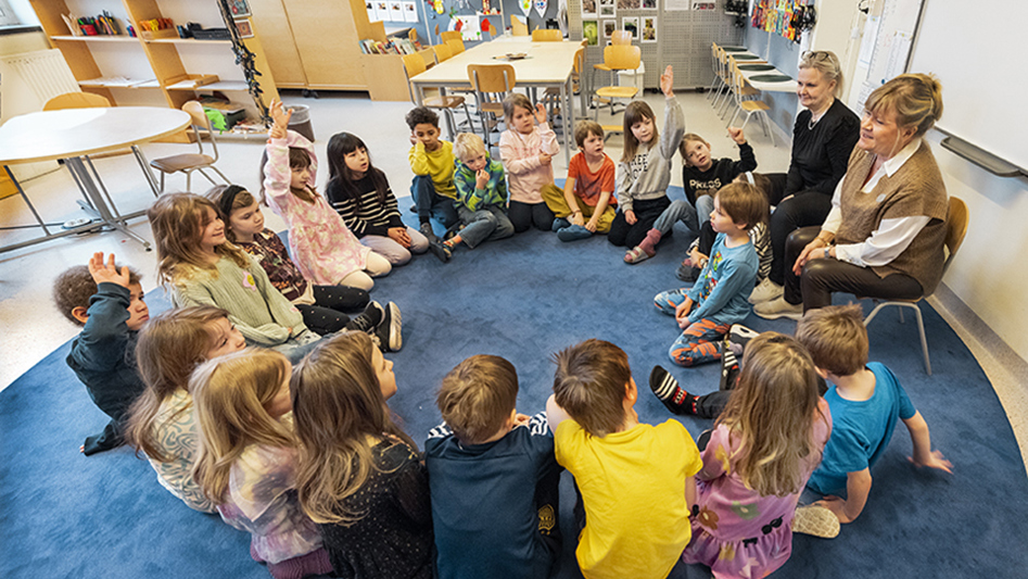 En grupp med barn och lärare sitter i en ring på golvet i ett klassrum.
