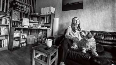 Kvinna med hund sitter i soffa i vardagsrum med bokhyllor runtomkring. Svartvit bild.