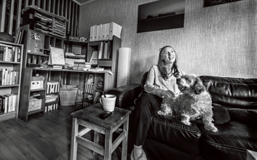 Kvinna med hund sitter i soffa i vardagsrum med bokhyllor runtomkring. Svartvit bild.