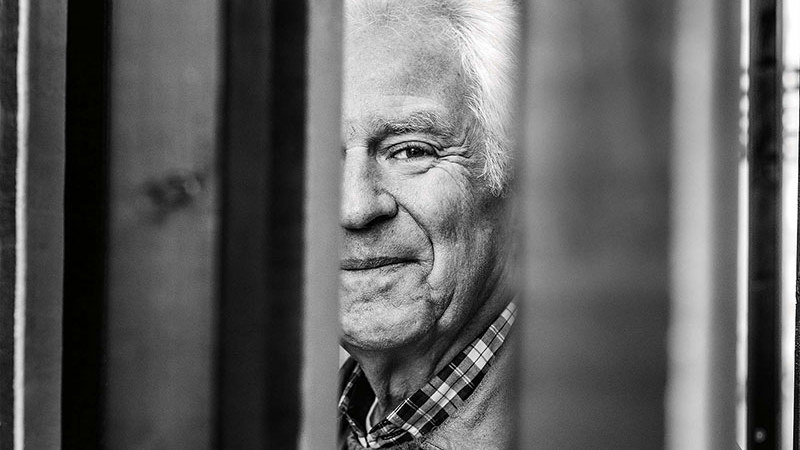 Närbild i svartvitt på äldre man som kikar fram bakom en dörr. Halva ansiktet syns.