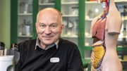 En man sitter i ett klassrum intill en anatomisk modell av en människokropp.