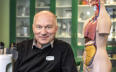 En man sitter i ett klassrum intill en anatomisk modell av en människokropp.