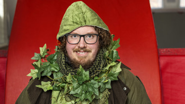 En leende man iklädd grön jacka med luva och med murgröna virad runt halsen.