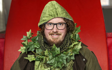 En leende man iklädd grön jacka med luva och med murgröna virad runt halsen.