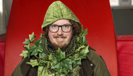 En leende man iklädd grön jacka med luva och med murgröna virad runt halsen.