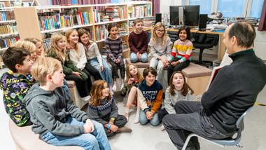 En man visar en uppslagen bok för elever som sitter framför honom på golvet.