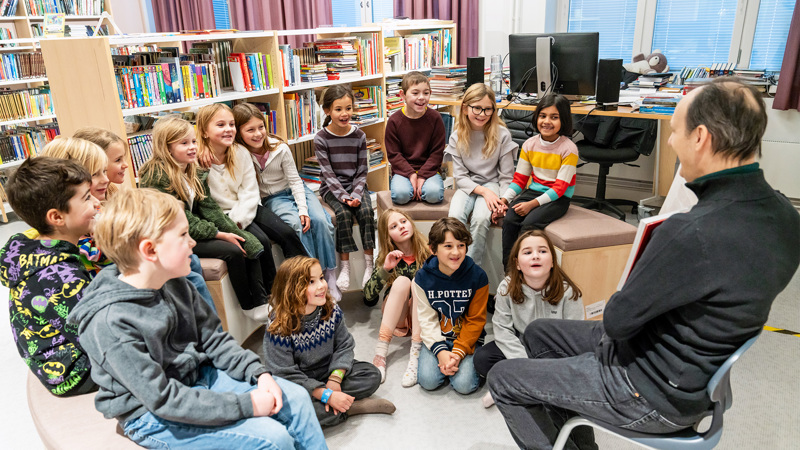 En man visar en uppslagen bok för elever som sitter framför honom på golvet.