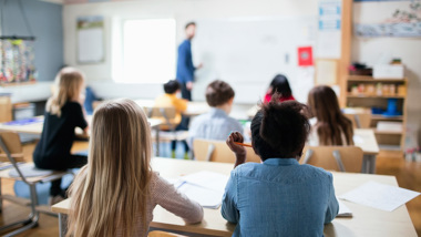 Barn i som tittar på läraren som står längst fram i klassrummet.