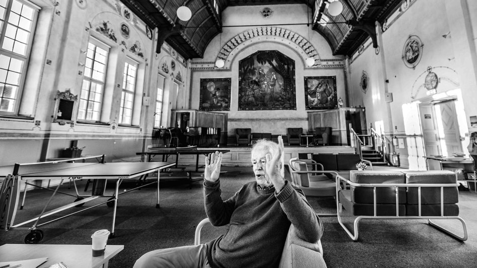 Äldre man som sitter i en rymlig byggnad med högt i tak. Han gestikulerar engagerat med händerna.