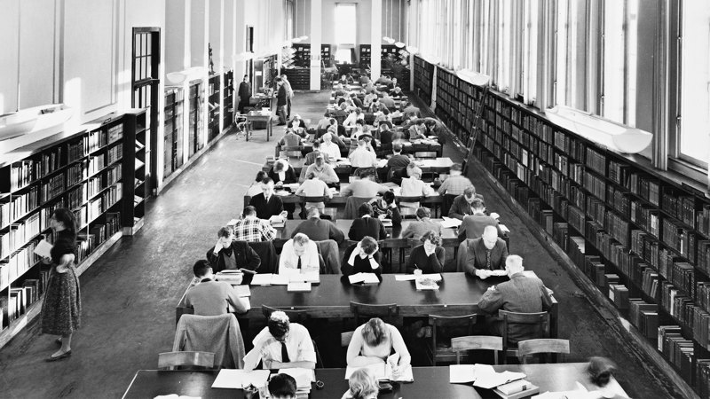 Gammalt svartvitt foto av människor som sitter och läser vid bord i ett bibliotek.