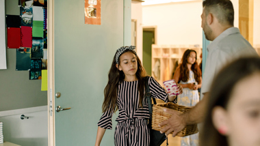 Barn går in i klassrum och lägger mobiltelefon i korg som lärare håller i.