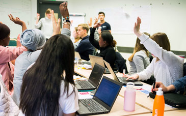 Elever sitter runt ett bord i ett klassrum, några med datorer framför sig, och räcker upp handen.