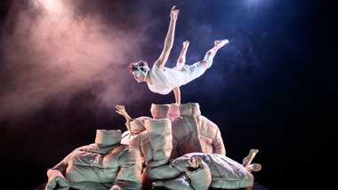 En dansare balanserar horisontellt på en hand ovanpå en hög med jackor.