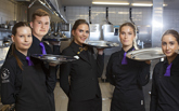 Fem servitörer står i ett restaurangkök och håller i varsin serveringsbricka
