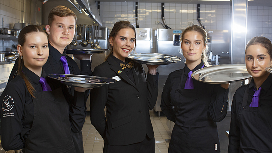 Fem servitörer står i ett restaurangkök och håller i varsin serveringsbricka
