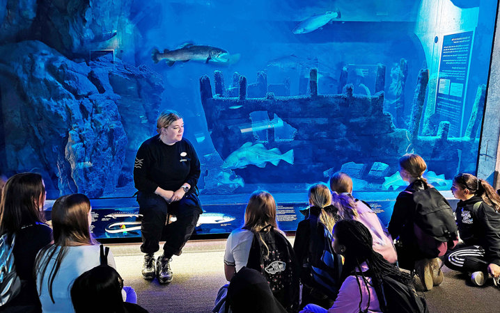 Kvinna sitter omgiven av barn framför stort akvarium med fiskar och skeppsvrak.