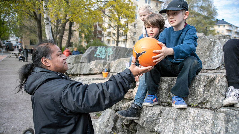 En pedagog ger en boll till några barn som sitter på en stor sten.