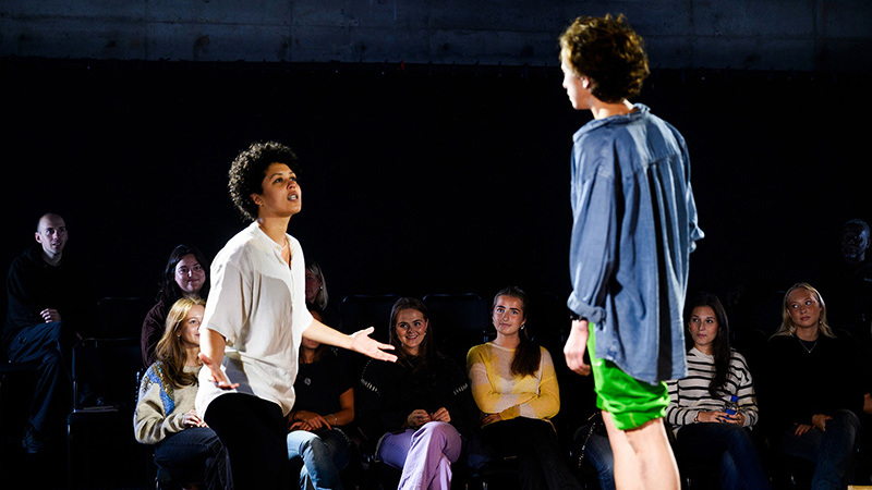 Två skådespelare interagerar på en scen i ett mörkt rum. I bakgrunden syns några ur publiken.
