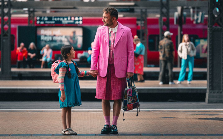 En flicka i blå klänning håller en man som är klädd i en rosa kavaj och kjol i handen.